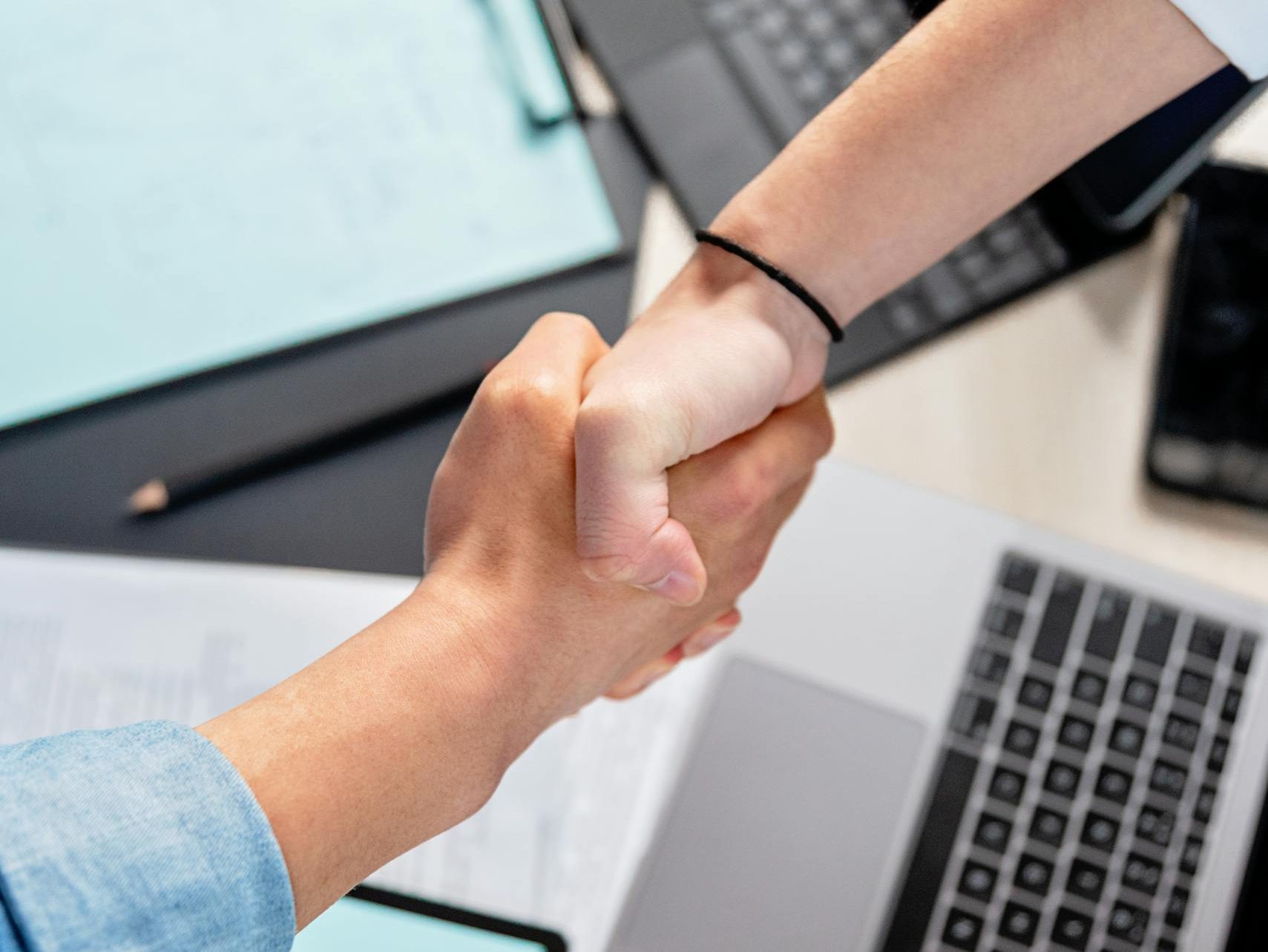 Two people shaking hands over laptops and documents in an office, symbolizing a business deal.