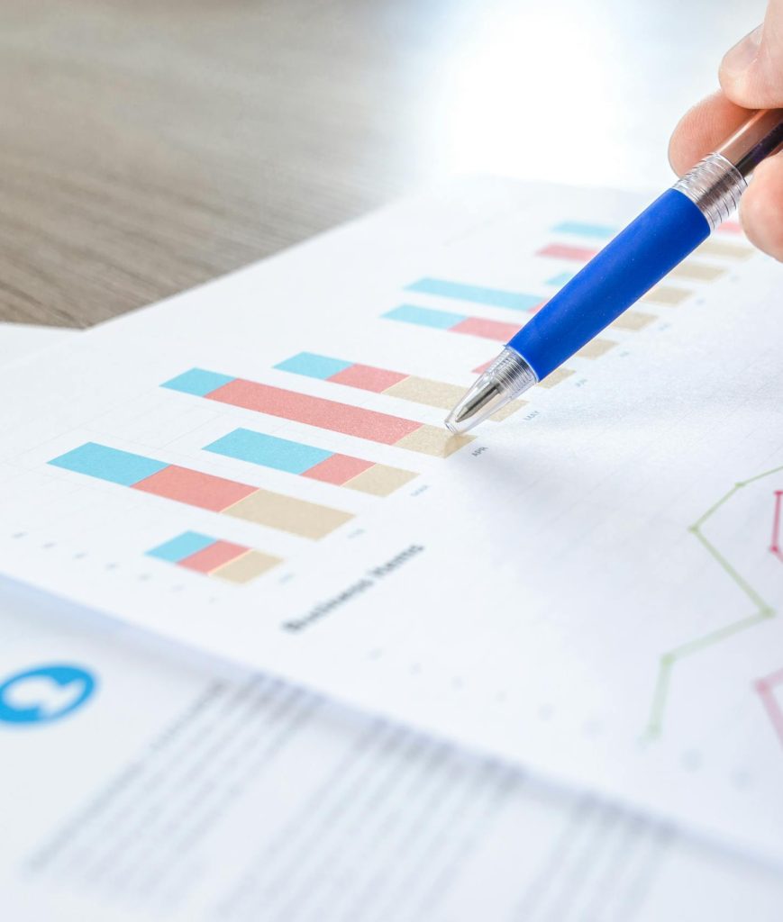 A close-up of a hand with a pen analyzing data on colorful bar and line charts on paper.